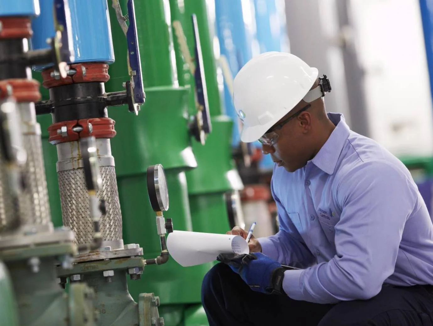 Mechanic wearing safety equipment recording readings beside building equipment